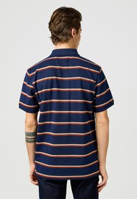 Kortärmad marinblå polo med horisontella ränder i orange och beige. Krage med rak fåll och en strukturerad tygblandning.