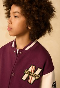 Bordeauxfarbene Collegejacke mit weißen Akzenten, versehen mit einem aufgestickten New York City-Patch. Druckknöpfe und ein gerippter Kragen runden das Design ab.