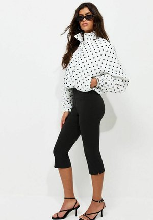 Vrouw draagt zwarte cropped broek en witte top met lange mouwen en zwarte polkadots, zwarte sandalen met hak en donkere zonnebril.