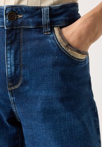 Jeans in denim di colore blu scuro con una finitura testurizzata, caratterizzati da cuciture dorate a contrasto e una tasca frontale con un'originale chiusura a zip.