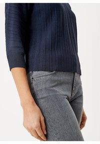 Gros plan sur une personne portant un pull côtelé bleu marine à manches trois-quarts et un jean gris délavé, debout devant un fond uni.