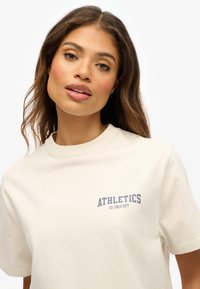 T-shirt en coton couleur crème avec col rond, arborant "ATHLETICS" en texte bleu marine sur la poitrine gauche. Manches courtes, texture lisse.