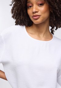 Giovane donna con capelli ricci che indossa un top bianco testurizzato, trucco leggero e un piccolo orecchino d'oro su uno sfondo semplice.