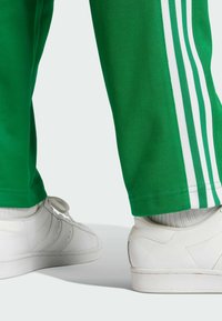 Pantalones deportivos verdes con rayas blancas en los lados, combinados con zapatillas blancas y calcetines grises. Vista en primer plano desde abajo.