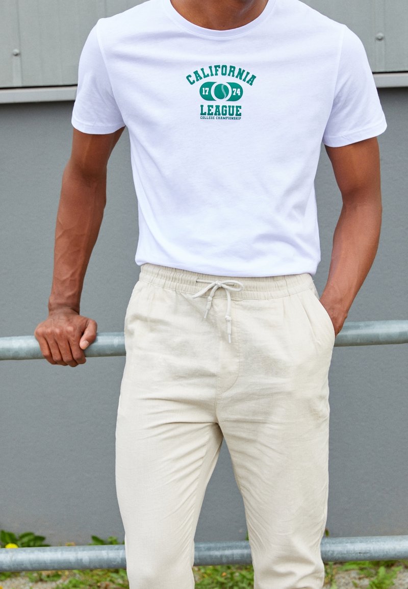 Homme portant un t-shirt blanc « California League 1774 College Championship » et un pantalon beige clair à cordon, appuyé sur une rambarde métallique à l'extérieur.