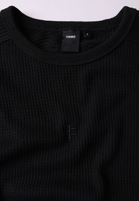 Camisola preta de malha canelada com decote redondo. Um pequeno logótipo bordado na frente adiciona textura ao design. Etiqueta de tamanho: L.