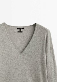 Pull à col en V à manches longues de couleur gris clair, en tissu doux avec une texture lisse. Étiquette visible au niveau du cou, portant la mention "Fabriqué en Chine."