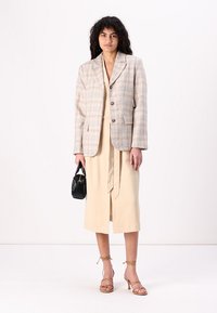 Blazer à carreaux beige avec deux boutons, associé à une robe beige avec une taille nouée. Accessoirisé avec un petit sac à main noir et des sandales à talons.