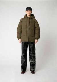 Olivgrüne gepolsterte Jacke mit Kapuze, elastischen Bündchen und Reißverschluss, kombiniert mit glänzenden schwarzen Vinylhosen und braunen Blockabsatzstiefeln.