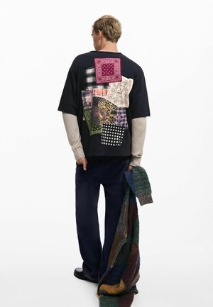 Black oversized t-shirt with a patchwork design on the back, featuring various fabrics and patterns. Model wearing blue pants and holding a multicolored scarf.