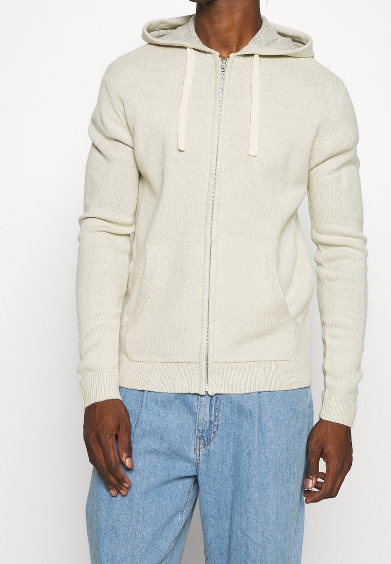 Felpa con zip di colore beige chiaro realizzata in tessuto a maglia testurizzato, dotata di cappuccio con cordino e tasche laterali. I polsini e l'orlo sono a costine.