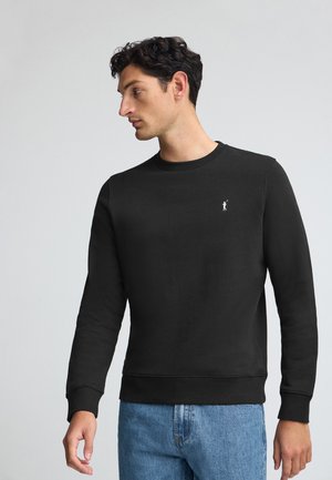 Black sweatshirt with a round neck, ribbed cuffs and hem, featuring a small logo on the chest. Worn with light blue denim jeans.