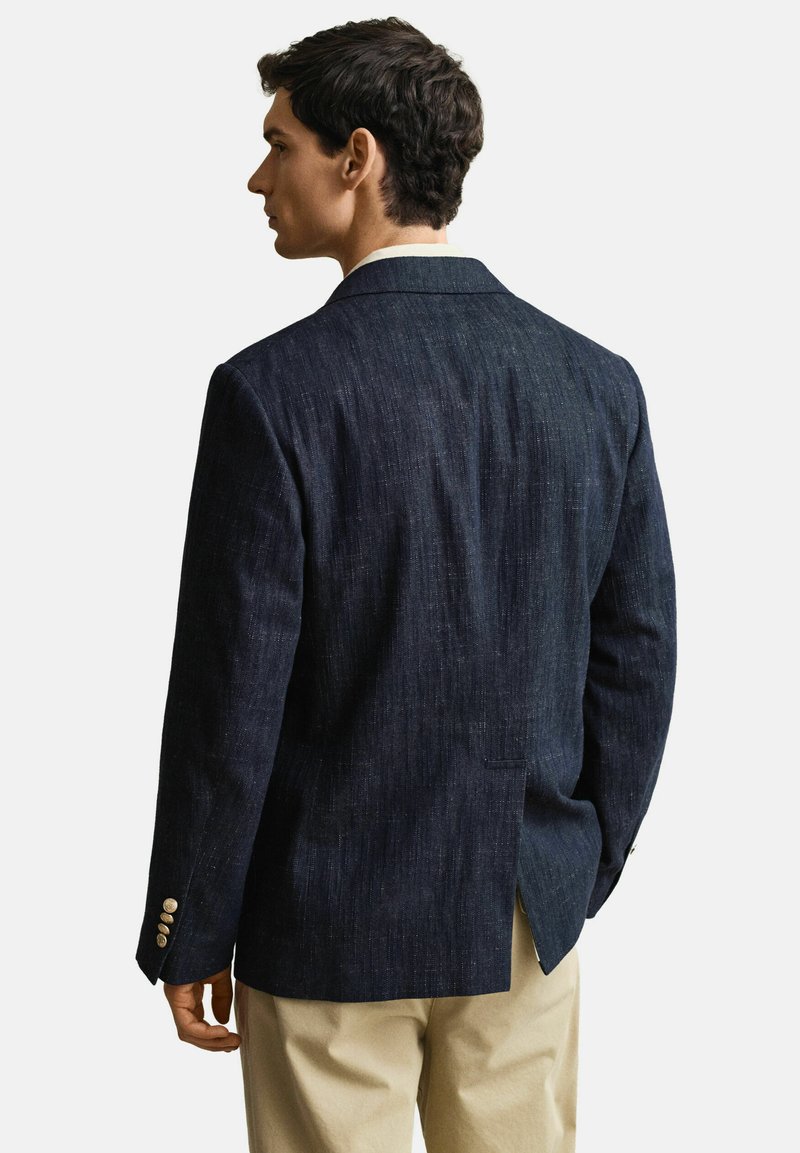 Mann trägt dunkelblauen Blazer mit goldenen Knöpfen und beigefarbene Hose, steht mit dem Rücken zu einem schlichten weißen Hintergrund.