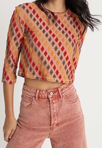 Femme portant un pull court à manches trois-quarts avec un motif losange multicolore et un jean rose délavé taille haute.