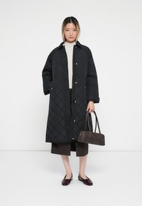 Black quilted coat with snap buttons, worn over a light sweater and dark pants. Holding a small dark handbag, with dark flat shoes.
