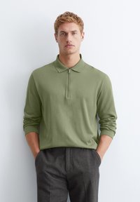 Olivengrüner Strickpullover mit einem Reißverschlusskragen und langen Ärmeln, kombiniert mit dunkelgrauen Hosen. Glatte Textur, lässige Passform.
