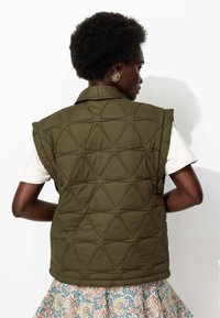 Gilet matelassé vert olive avec un motif triangulaire texturé, doté d'un col et sans manches, porté sur une chemise blanche et une jupe à motifs.