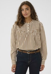 Donna con capelli ricci indossa una blusa a maniche lunghe a righe beige e bianche infilata in jeans scuri con una cintura marrone, in piedi di fronte a uno sfondo bianco.