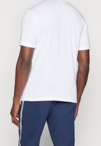 Man med en enkel vit T-shirt med kort ärm och blå träningsbyxor med vit rand och textdetaljer på sidan, stående med ryggen vänd.