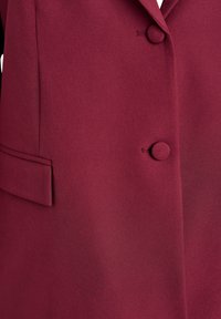 Nærbillede af en burgunder blazer, der viser stofbetrukne knapper og en klaplomme foran.