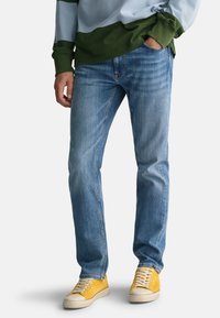 Light blue jeans with a straight fit, featuring subtle fading. Paired with yellow suede sneakers. Green and blue striped sweater visible above.