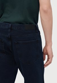 Gros plan d'une personne portant un jean en denim bleu foncé et une chemise vert foncé, montrant la poche arrière et une partie du bras droit pendant.