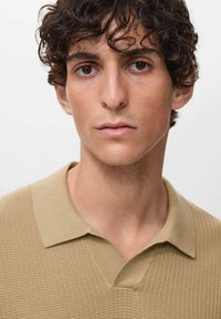Beige ribbed polo shirt with a short collar, textured fabric, fitted design, and subtle stitching details, worn by a model with curly hair.