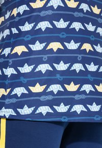 Modrá látka s opakujícím se vzorem origami lodí v světle modré, žluté a bílé barvě, prokládaných modrými provazy.