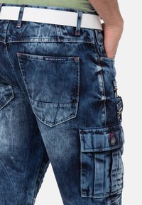 Cargo-Jeans aus Denim in einem distressed Dunkelblau mit mehreren Taschen, roten Nähdetails und einem weißen Gürtel.