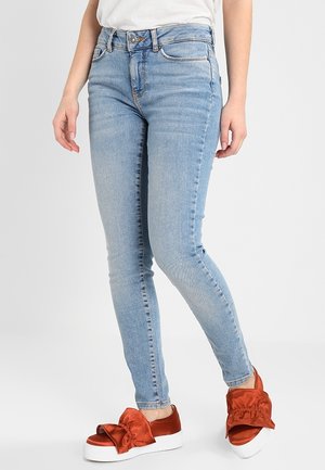 Personne portant un jean skinny bleu clair et des chaussures slip-on rouges avec de grands nœuds sur des semelles plateformes blanches.