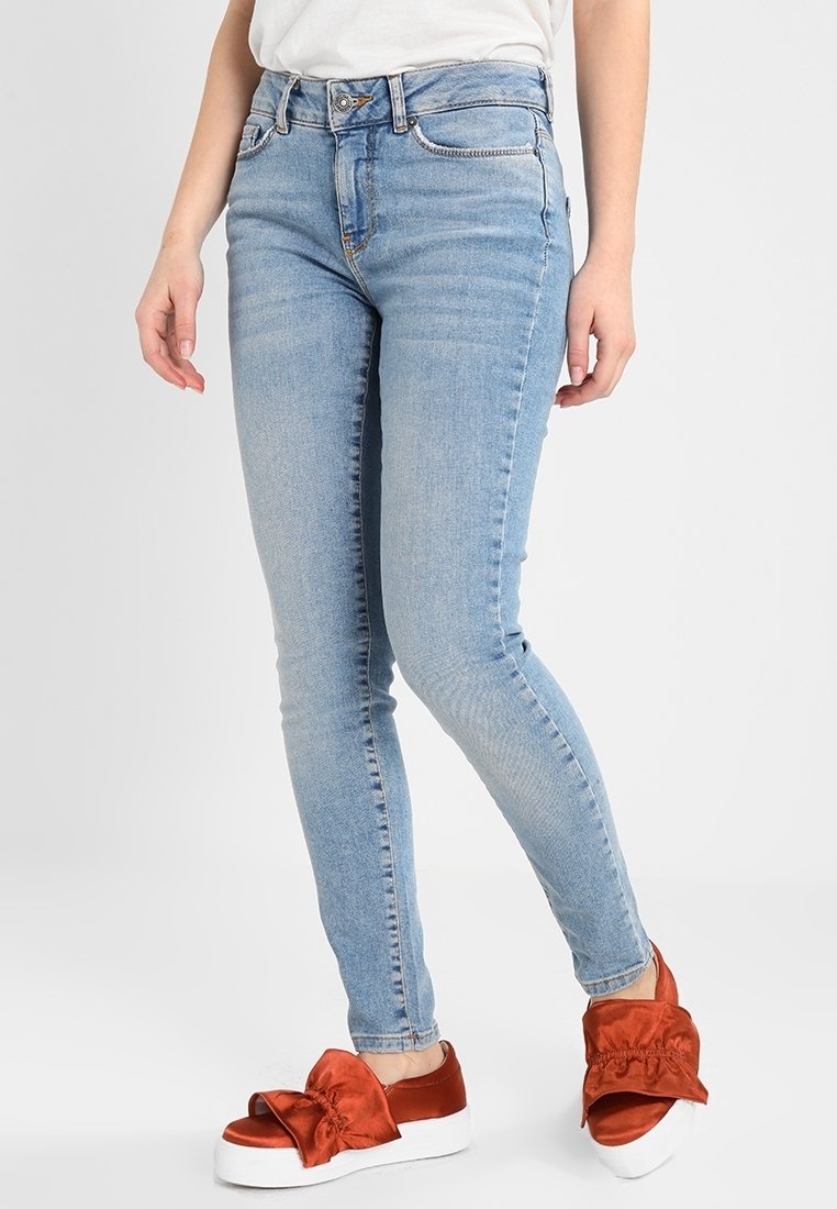 Personne portant un jean skinny bleu clair et des chaussures slip-on rouges avec de grands nœuds sur des semelles plateformes blanches.