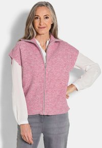 Gilet tricot rose à côtes avec un col haut et une fermeture éclair à l'avant. Manches courtes, texture douce et coupe décontractée. Porté sur une blouse blanche.