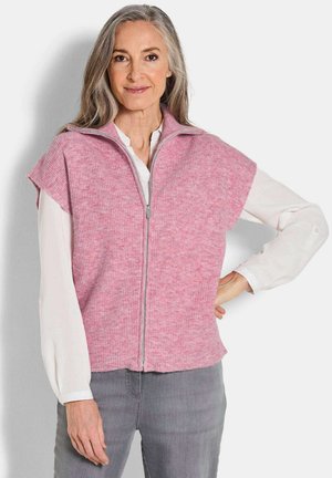 Rosa, gerippter Strickwesten mit hohem Kragen und Reißverschluss vorne. Mit kurzen Ärmeln, weichem Material und lockerer Passform. Getragen über einer weißen Bluse.