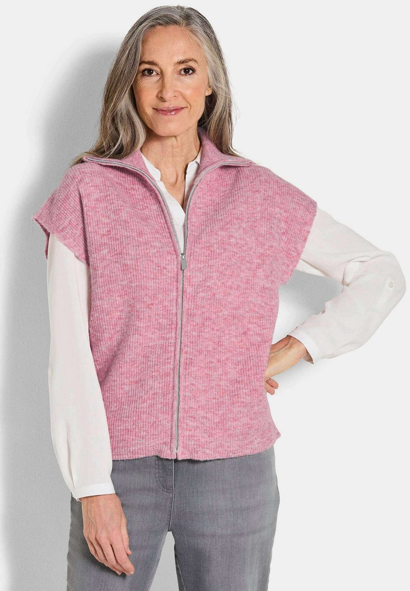 Gilet tricot rose à côtes avec un col haut et une fermeture éclair à l'avant. Manches courtes, texture douce et coupe décontractée. Porté sur une blouse blanche.