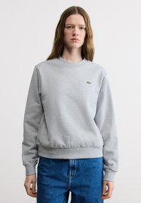 Jeune femme aux longs cheveux bruns portant un sweat-shirt gris clair avec un petit logo de crocodile vert et un jean bleu, debout devant un fond blanc.