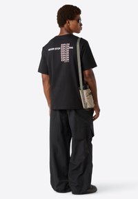 Man som bär en svart t-shirt med texten "Never Stop Exploring", svarta byxor, solglasögon och bär en beige axelväska med en vattenflaska.