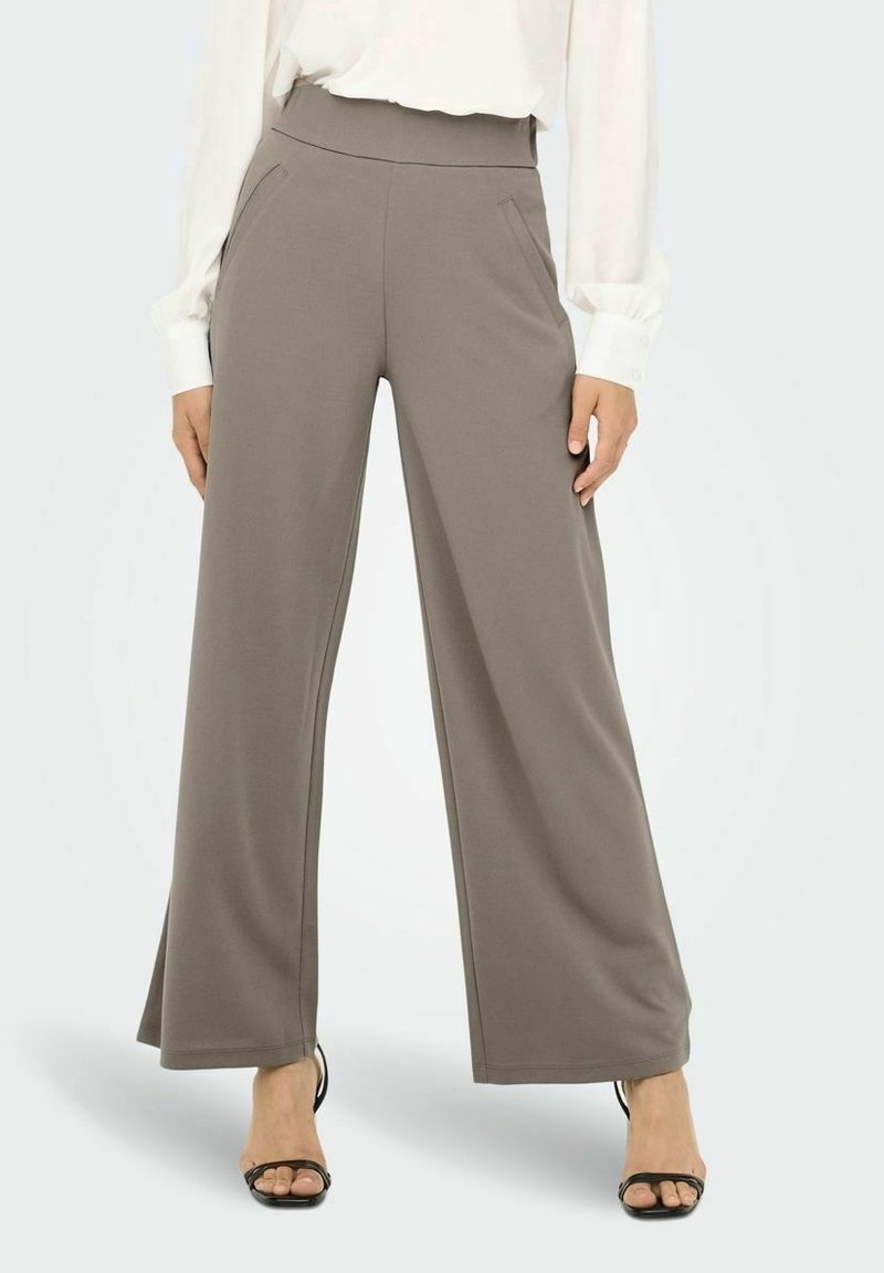 Femme portant un pantalon large taupe avec poches latérales, associé à un chemisier blanc à manches longues et des talons noirs ouverts à l’avant.