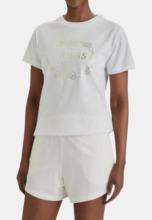 Persona che indossa una t-shirt bianca a maniche corte con il logo dorato "GUESS" e pantaloncini bianchi davanti a uno sfondo chiaro uniforme.