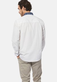 Weißes Langarmhemd mit einem gemusterten blauen Kragen, mit gewelltem Saum, Knopfmanschetten und einer glatten Textur. Getragen mit hellbeigen Hosen.