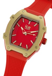 Reloj de silicona rojo con una caja hexagonal en tono dorado, que presenta un atrevido dial rojo, marcadores de hora dorados y una correa texturizada.