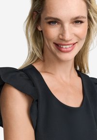 Vestido negro con mangas acampanadas, escote redondo y textura de tela suave. La modelo tiene el cabello rubio claro y un look de maquillaje natural.