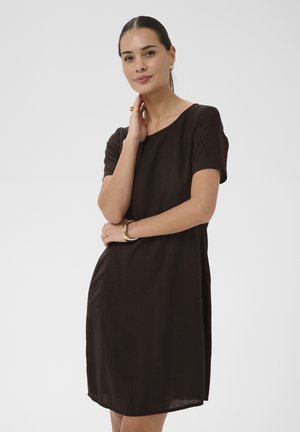 Kvinde med mørkt hår i en lav hestehale iført en simpel, kortærmet sort kjole, guldringeøreringe, armbånd og ring, poserende med hånden nær halsen.