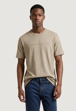 Giovane uomo che indossa una t-shirt beige "Armani Exchange" e jeans blu, in piedi davanti a uno sfondo chiaro e uniforme.