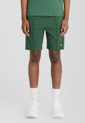 Mand iført grønne shorts med gul logo på venstre ben, hvide sokker og hvide atletik sko, stående mod en ensfarvet baggrund.
