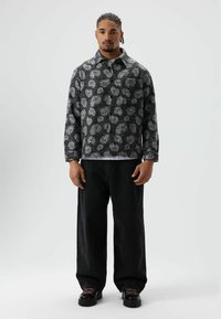 Zwart-grijs patroon shirtjack met een kraag, een borstzak en lange mouwen, gecombineerd met losse zwarte jeans en zwarte schoenen.