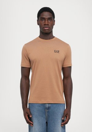 Brown short-sleeve t-shirt made of soft fabric, featuring a round neck and a small black logo on the chest. Worn with light blue jeans.