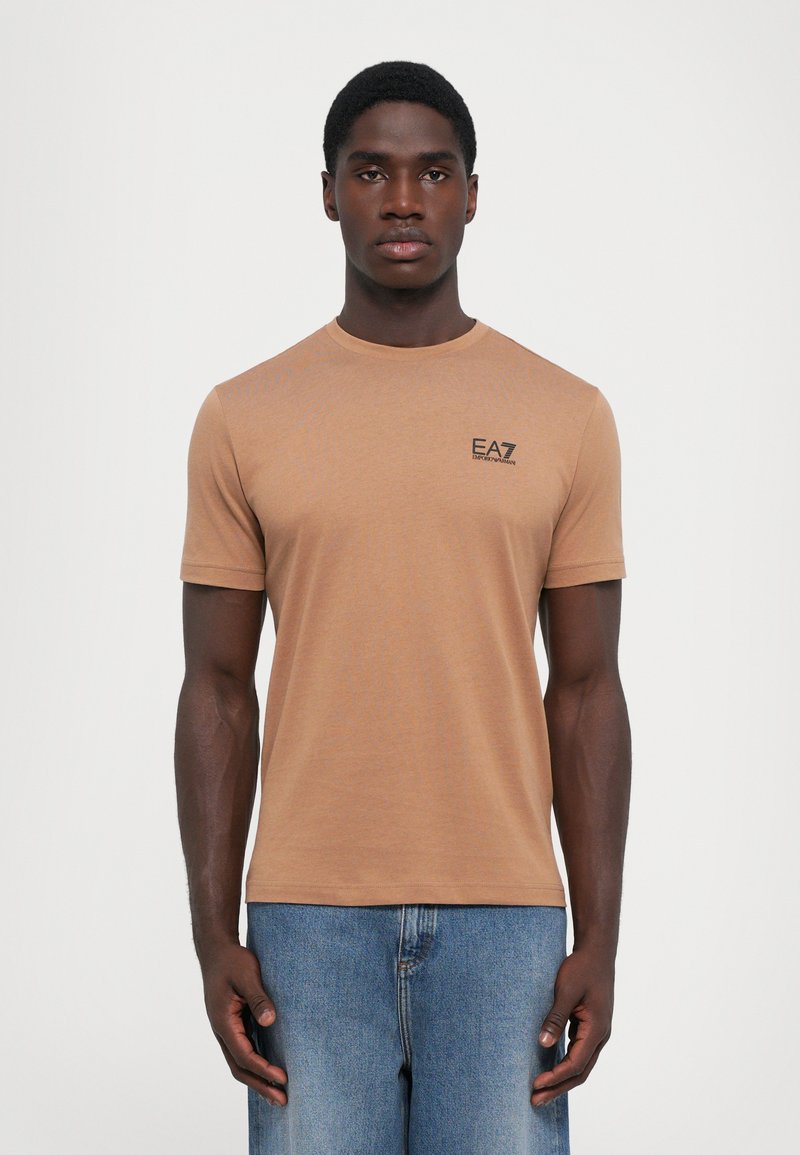 Brun t-skjorte med korte ermer laget av mykt stoff, med rund hals og en liten svart logo på brystet. Båret med lyseblå jeans.