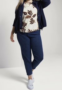 Top floreale bianco con motivo a foglie scure, giacca blu navy, jeans blu e sneaker bianche. L'outfit presenta una vestibilità comoda e un design informale.