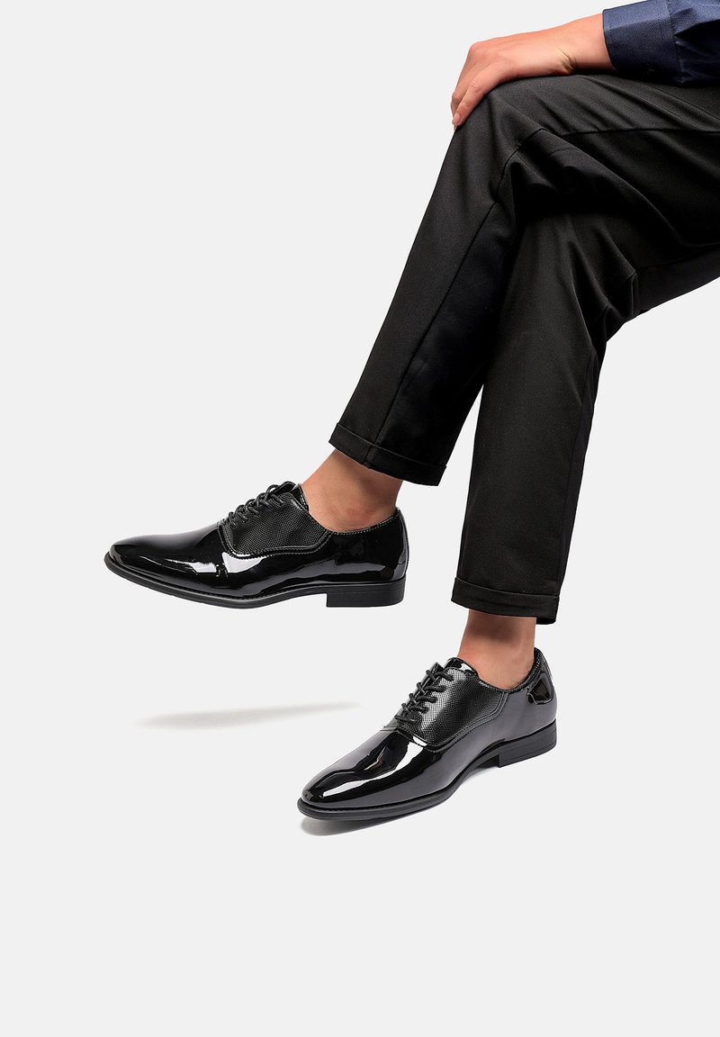 Scarpe eleganti in pelle nera con finitura lucida, design con lacci e accenti testurizzati, abbinate a un paio di pantaloni neri.