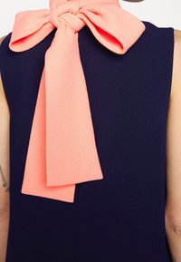 Personne portant un haut sans manches bleu marine foncé avec un grand nœud rose doux noué à l'arrière du cou, bras visibles avec des tatouages.